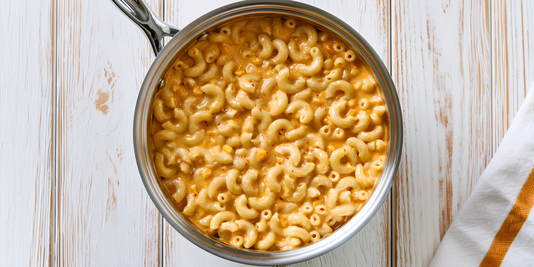 Overhead view of a stainless steel saucepan filled with spicy nacho cheese mac and cheese, with elbow pasta coated in a thick creamy orange sauce flecked with seasoning, set on a whitewashed wooden surface beside a white towel with a mustard-yellow stripe.